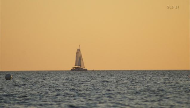 Ein Segelboot auf dem Meer im Sonnenuntergang. Der Himmel leuchtet gelb. Auf dem Wasser schwimmt im linken vorderen Bildbereich eine Boje.