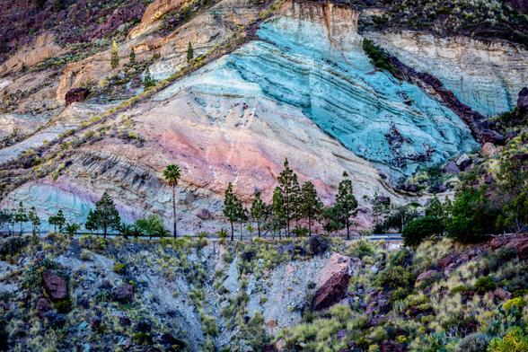 Dieses Foto zeigt eine atemberaubende Berglandschaft mit auffälligen, bunten Gesteinsschichten. Die Hänge erstrahlen in sanften Pastellfarben von hellem Blau über zartes Rosa bis hin zu warmen Beigetönen. Entlang des Berghangs verläuft eine Straße und davor stehen einige grüne Bäume und Palmen. Im Vordergrund erstreckt sich ein Hang mit spärlicher Vegetation und dunklen Steinen. Das natürliche Licht lässt die Farben des Gesteins besonders lebendig wirken und schafft eine eindrucksvolle Kulisse.