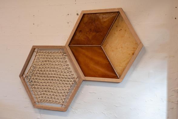 Two adjacent wooden hexagon frames hanging on a white wall; one containing a triaxial woven structure made of sisal rope; another segmented in 3 diamond-shape regions containing dried SCOBY biocellulose with different textures.